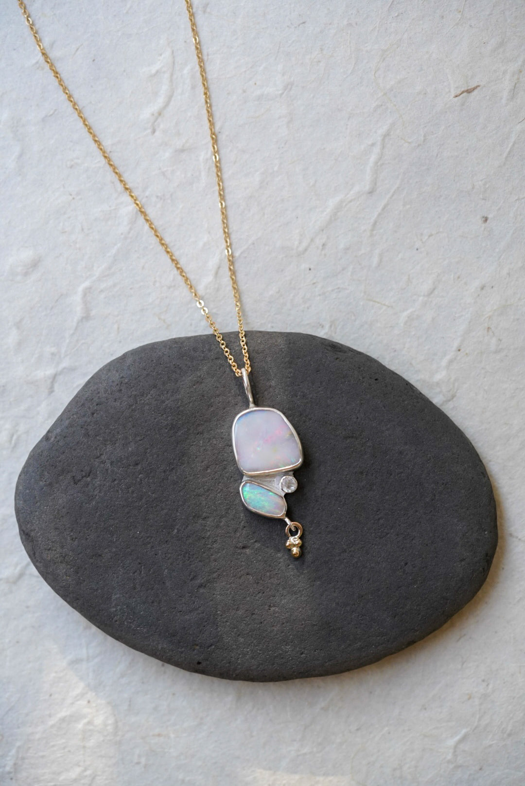 A gold chain necklace with a silver pendant featuring a large iridescent opal and a smaller green-blue opal opal, accented by a moonstone and a dangling gold bead, displayed on a dark gray stone against a light textured background.