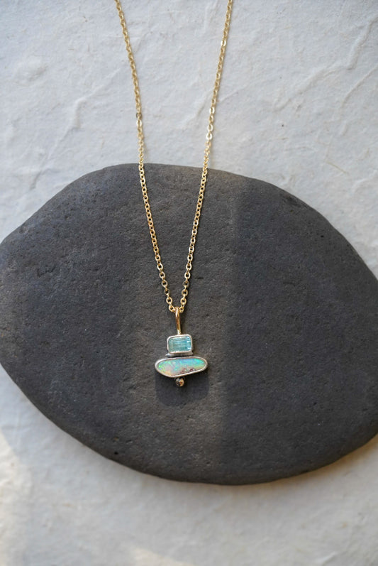 A delicate gold chain necklace with a pendant made of stacked natural stones, one tourmaline and one opal, displayed on a dark gray stone slab against a textured off-white background.