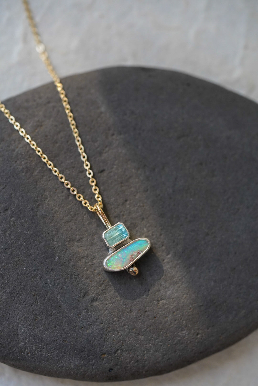 A delicate gold chain necklace with a pendant made of stacked natural stones, one tourmaline and one opal, displayed on a dark gray stone slab against a textured off-white background.