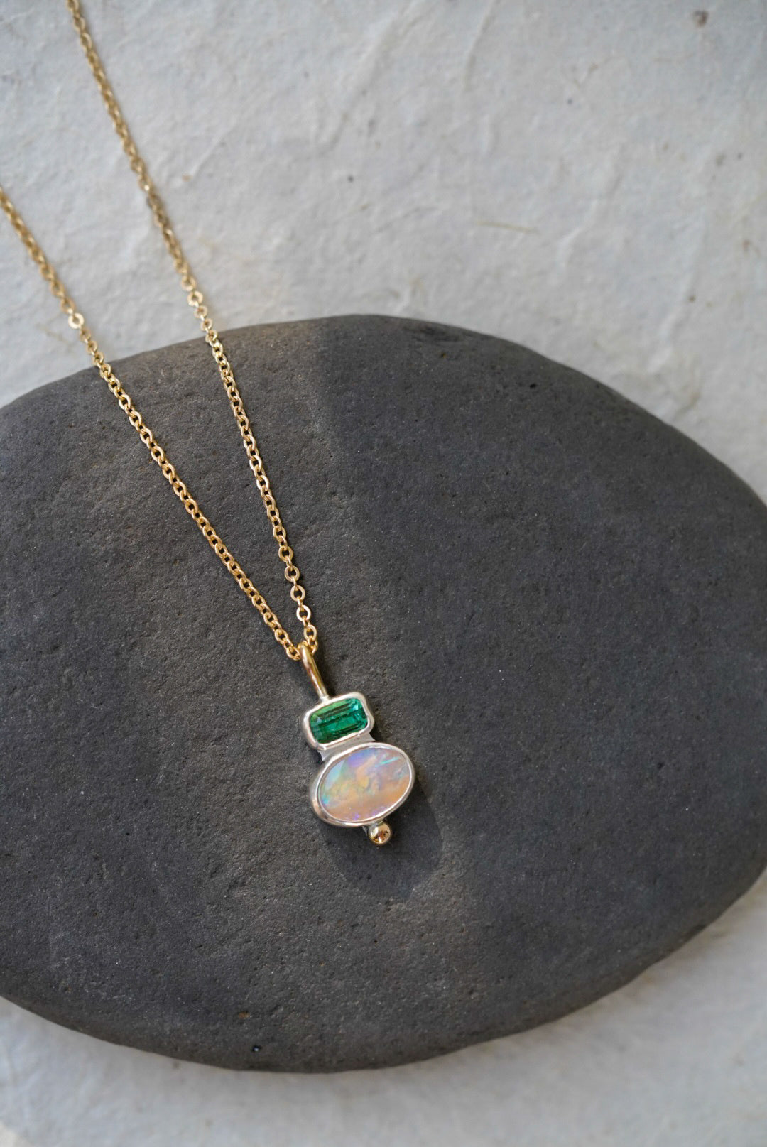 A delicate gold chain necklace with a silver pendant featuring a green rectangular tourmaline and an oval iridescent opal, displayed on a dark gray stone against a light gray background.