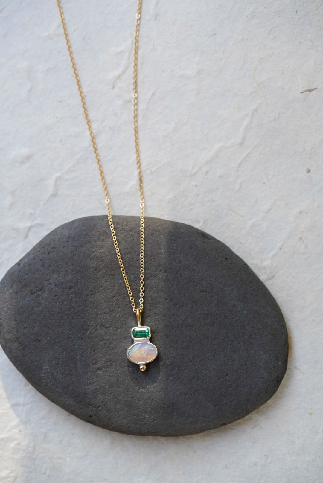 A delicate gold chain necklace with a silver pendant featuring a green rectangular tourmaline and an oval iridescent opal, displayed on a dark gray stone against a light gray background.