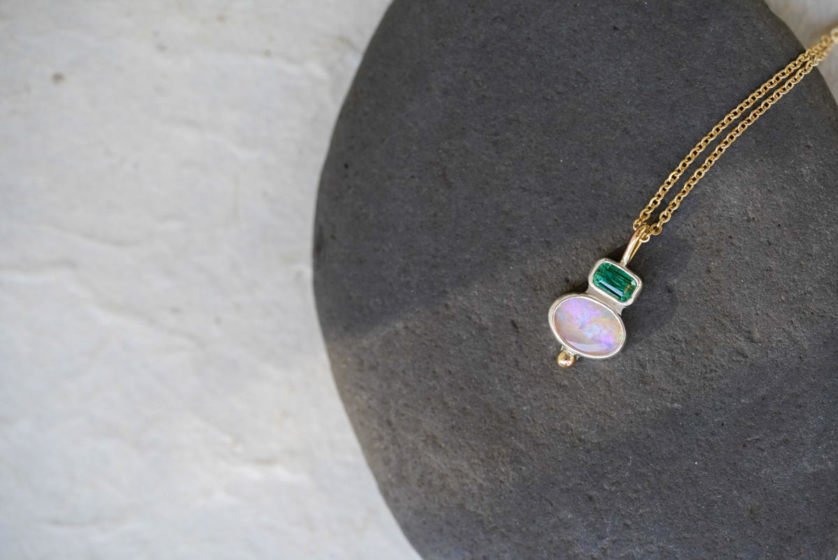 A delicate gold chain necklace with a silver pendant featuring a green rectangular tourmaline and an oval iridescent opal, displayed on a dark gray stone against a light gray background.