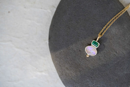 A delicate gold chain necklace with a silver pendant featuring a green rectangular tourmaline and an oval iridescent opal, displayed on a dark gray stone against a light gray background.