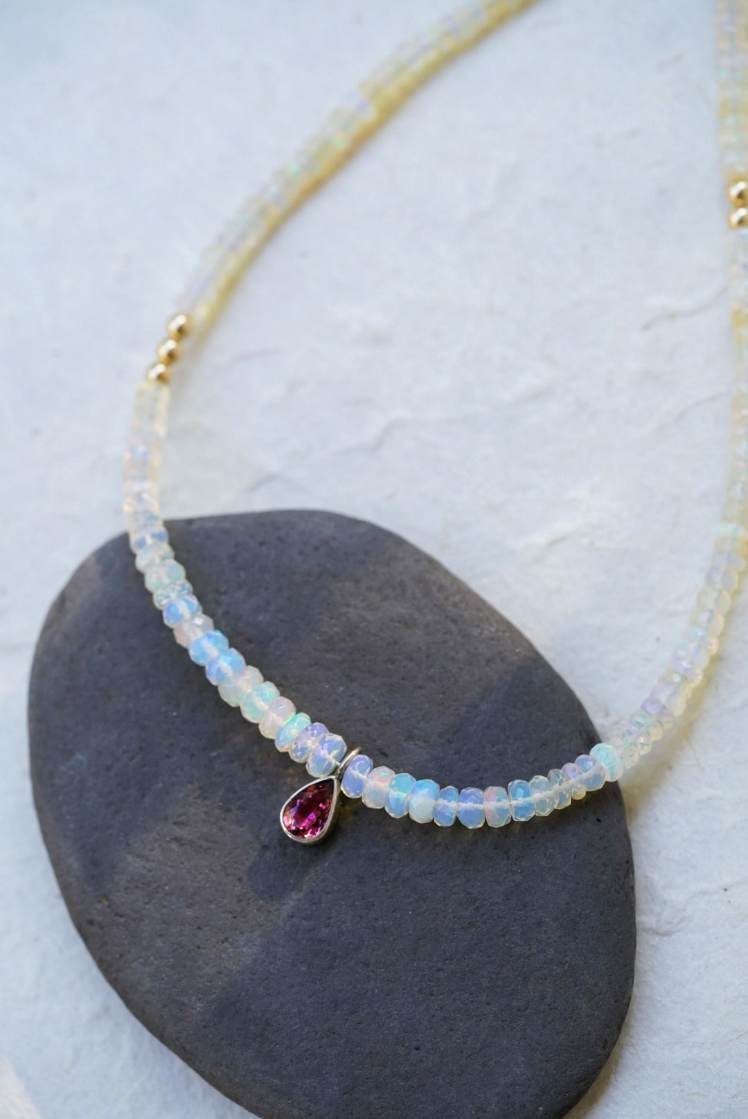 A opal strand beaded necklace accented with small gold beads and a dark pink teardrop tourmaline, displayed on a dark stone against a textured off-white background.