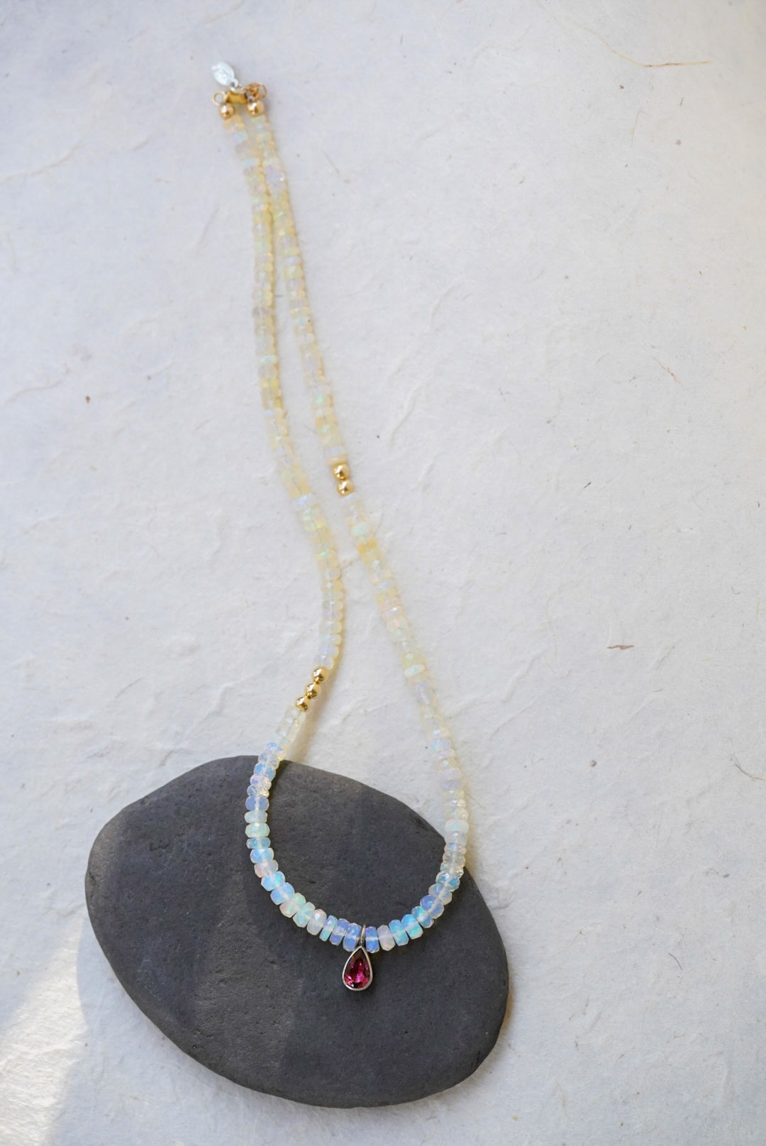 A opal strand beaded necklace accented with small gold beads and a dark pink teardrop tourmaline, displayed on a dark stone against a textured off-white background.