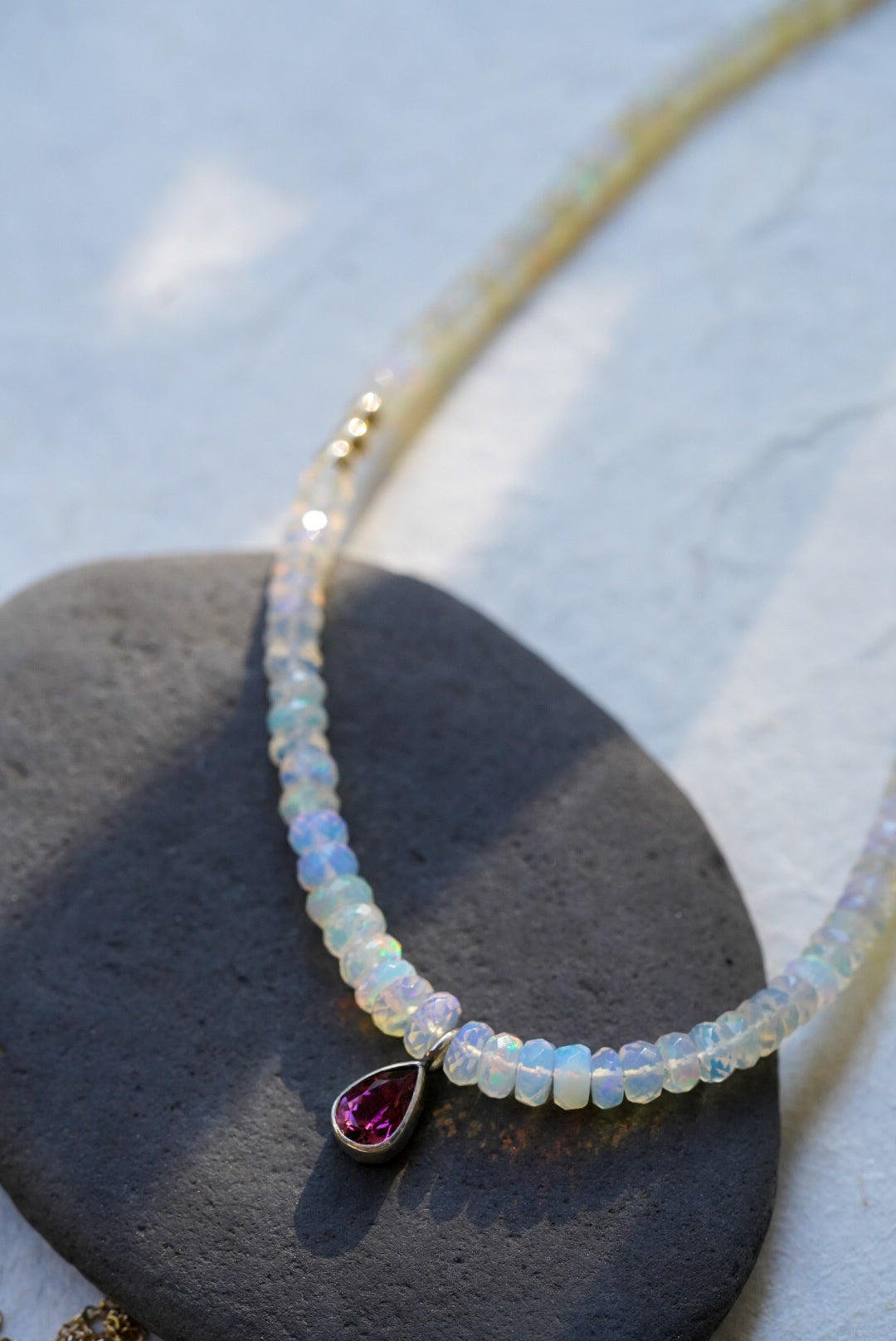 A opal strand beaded necklace accented with small gold beads and a dark pink teardrop tourmaline, displayed on a dark stone against a textured off-white background.