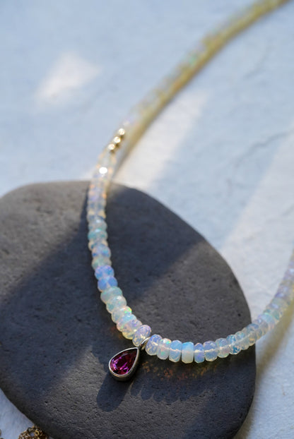 A opal strand beaded necklace accented with small gold beads and a dark pink teardrop tourmaline, displayed on a dark stone against a textured off-white background.