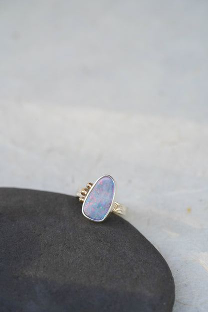 A silver ring with a teardrop-shaped iridescent opal gemstone with gold accents along the opal, resting on a dark textured stone against a light gray background.