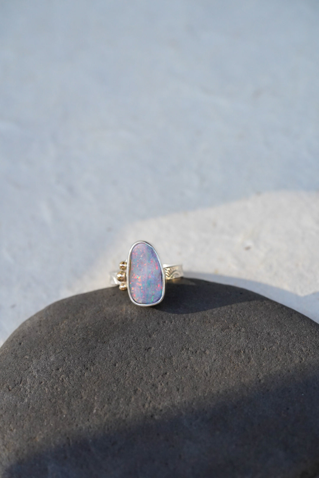 A silver ring with a teardrop-shaped iridescent opal gemstone with gold accents along the opal, resting on a dark textured stone against a light gray background.