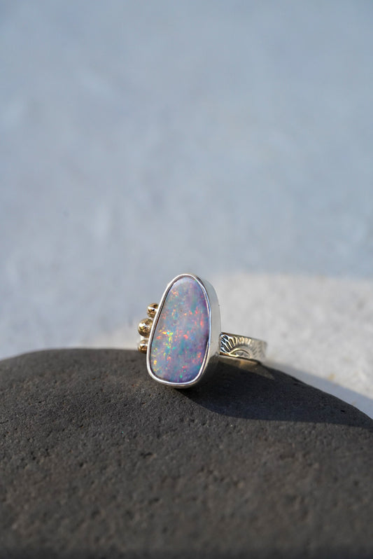 A silver and gold accented ring featuring a teardrop-shaped iridescent opal gemstone, resting on a dark textured stone against a soft, out-of-focus light gray background.