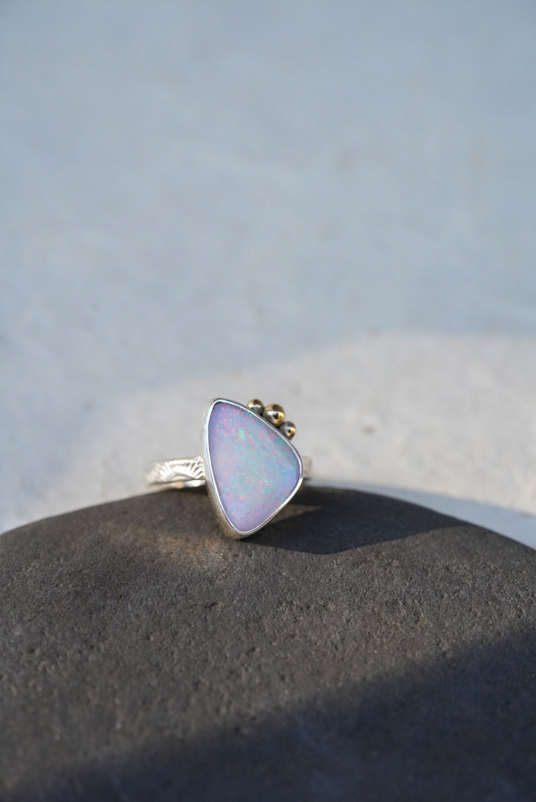 A triangular iridescent opal ring with a silver stamped band, resting on a dark textured stone against a light gray background.