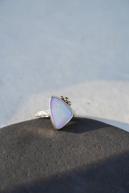 A triangular iridescent opal ring with a silver stamped band, resting on a dark textured stone against a light gray background.