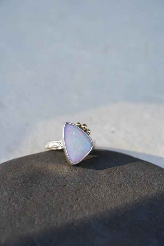 A triangular iridescent opal ring with a silver stamped band, resting on a dark textured stone against a light gray background.