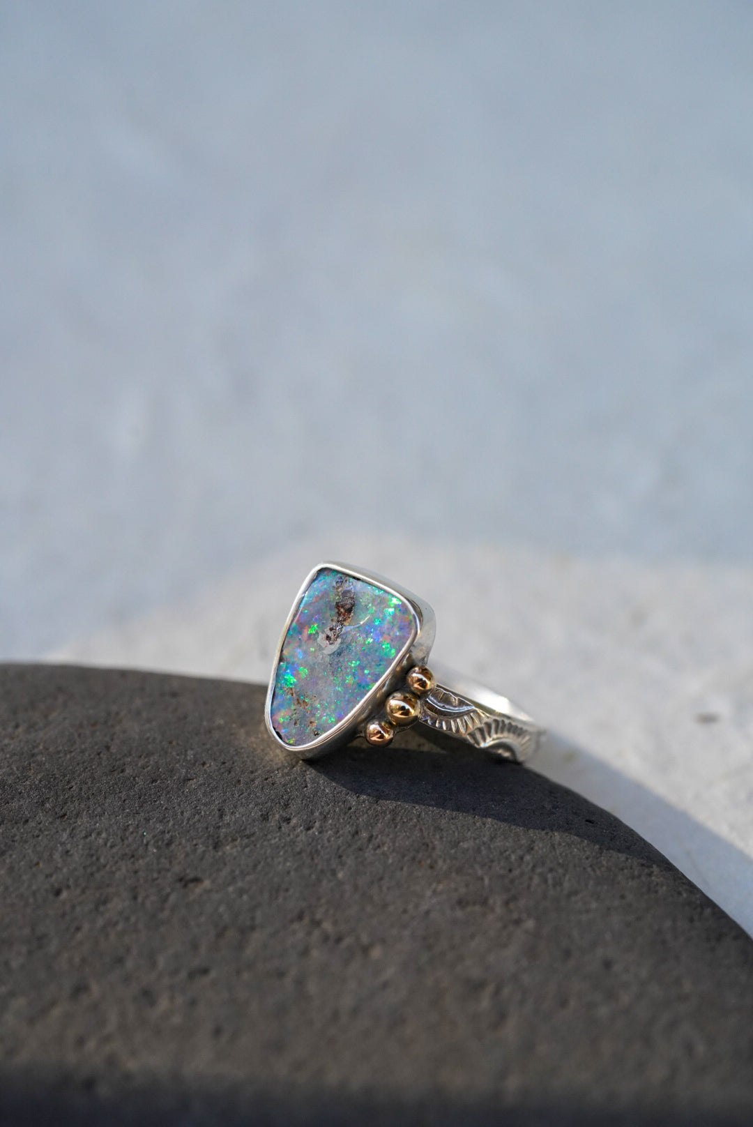 A silver ring with a triangular iridescent opal gemstone and small gold decorative accents on the band, resting on a dark textured surface against a light gray background.