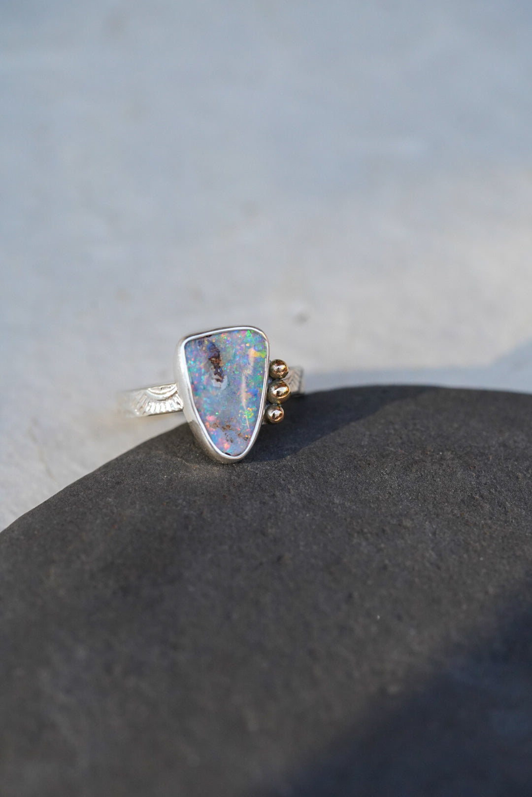 A silver ring with a triangular iridescent opal gemstone and small gold decorative accents on the band, resting on a dark textured surface against a light gray background.