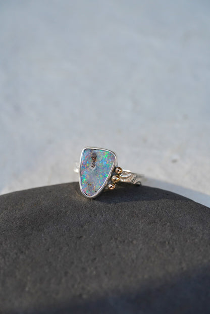 A silver ring with a triangular iridescent opal gemstone and small gold decorative accents on the band, resting on a dark textured surface against a light gray background.