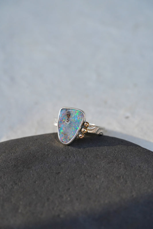 A silver ring with a triangular iridescent opal gemstone and small gold decorative accents on the band, resting on a dark textured surface against a light gray background.