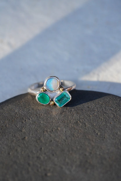 A silver ring with three gemstones—a rectangular emerald-cut emerald, a round emerald stone, and a round white opal—resting on a dark textured surface against a light gray background.