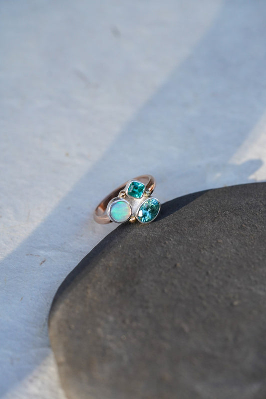 A rose gold ring with three green-blue gemstones, resting on a dark stone against a light gray background.