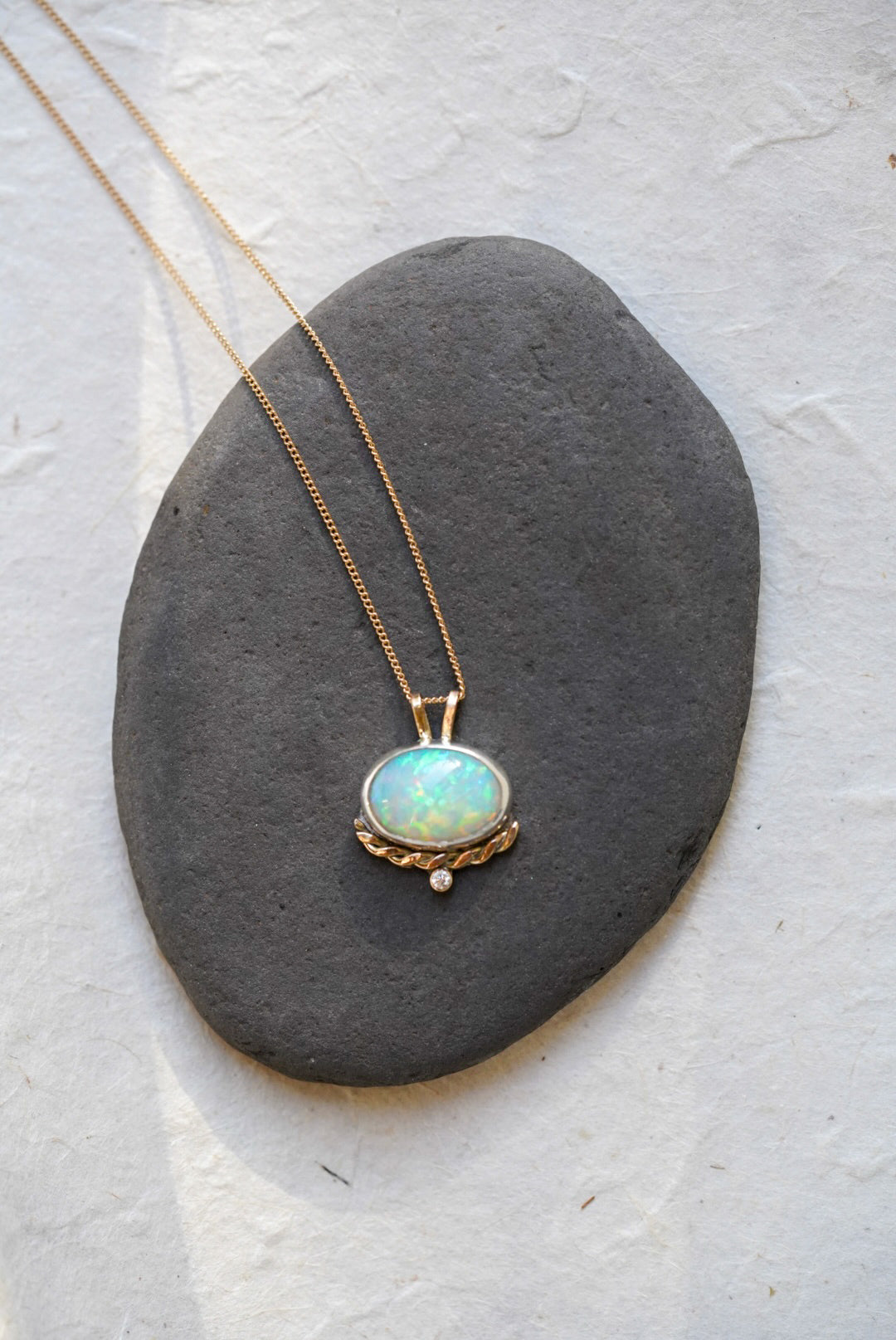 A gold necklace with an oval iridescent opal pendant with a twisted gold accent and small diamond below, resting on a dark gray stone against a textured off-white background.