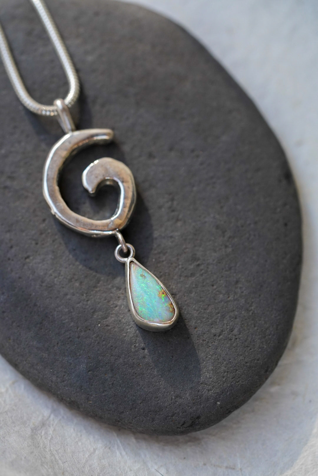 A silver necklace with a spiral pendant and a teardrop-shaped iridescent opal drop, displayed on a dark stone against a textured off-white background.