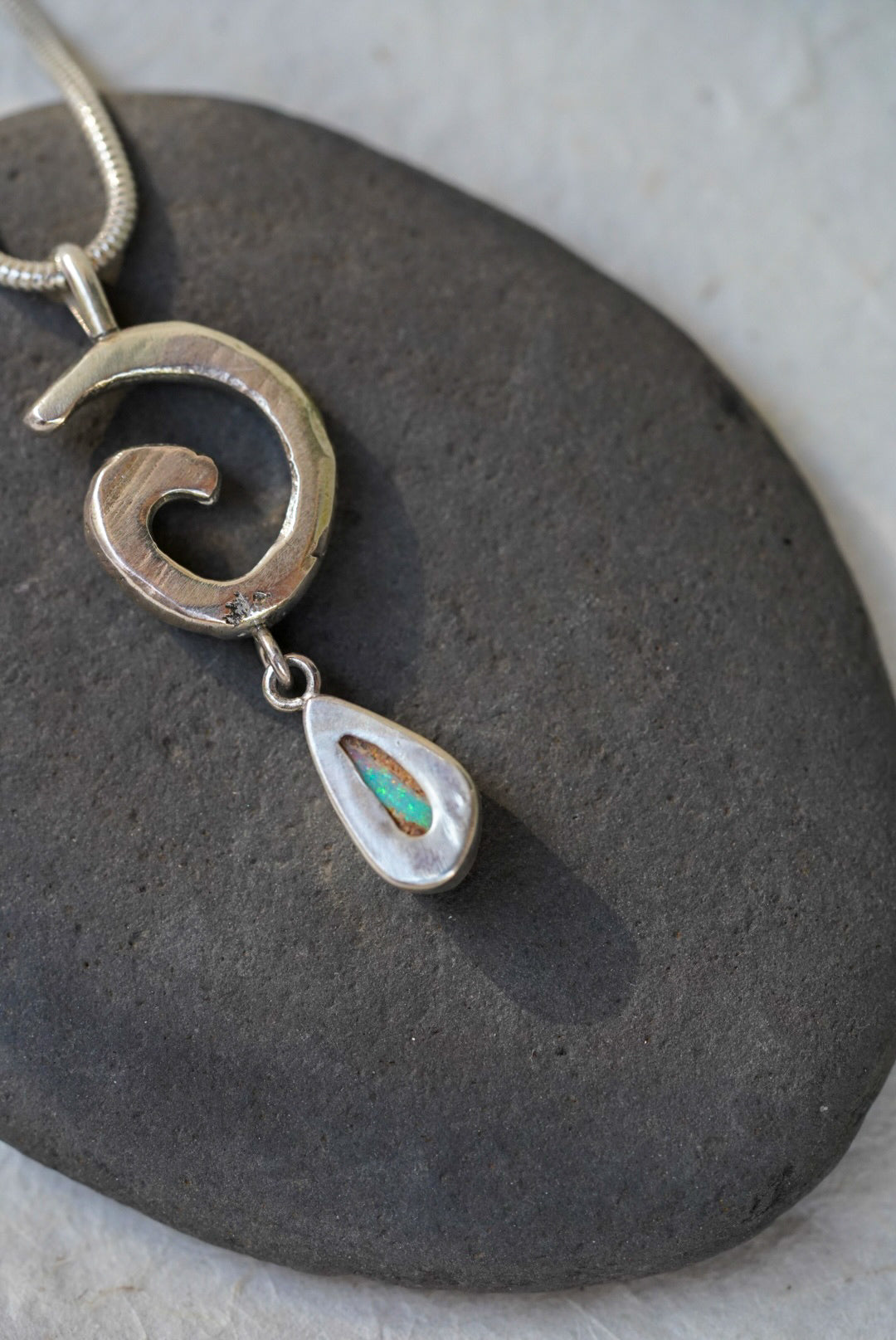 A silver pendant necklace with a textured spiral design and a dangling teardrop charm featuring a small green opening in the back, displayed on a dark gray stone.