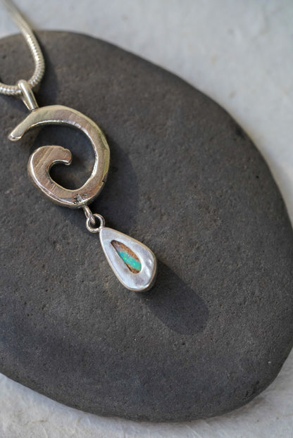 A silver pendant necklace with a textured spiral design and a dangling teardrop charm featuring a small green opening in the back, displayed on a dark gray stone.