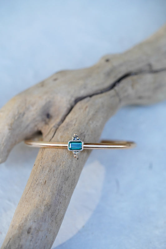 A delicate gold cuff featuring a small rectangular blue-green gemstone, displayed on a curved piece of driftwood against a light textured background.