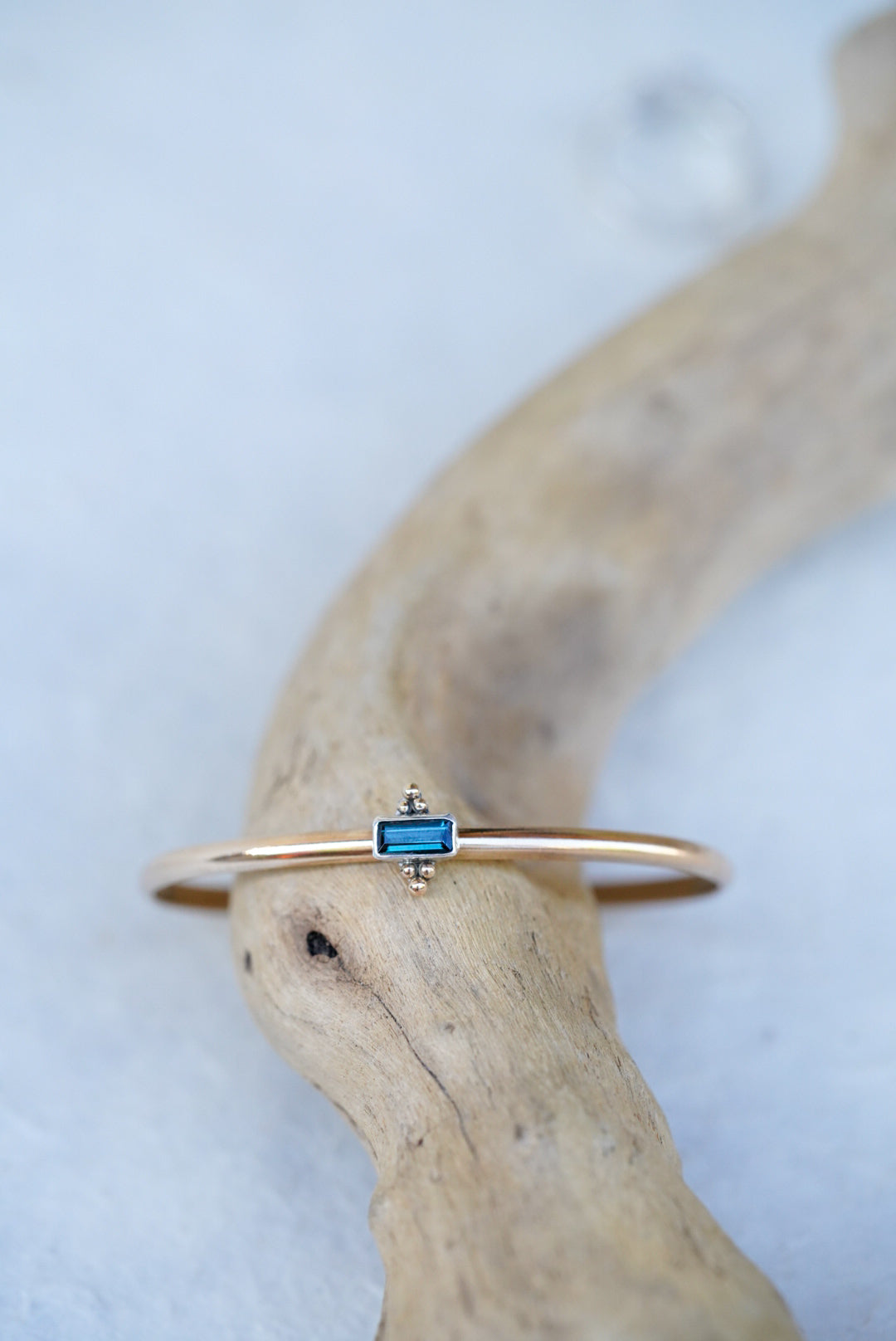 A delicate gold cuff featuring a small rectangular blue-green gemstone, displayed on a curved piece of driftwood against a light textured background.
