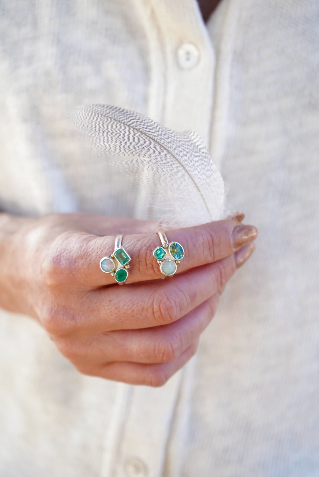 A close-up of a person's hand holding a delicate white feather, wearing two rings set with green and blue stones, against a soft cream-colored linen shirt.