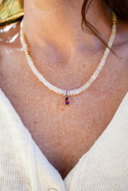 A close-up of someone wearing a opal beaded necklace with a few gold beads, at the center is a dark pink tourmaline, person is wearing a white knit top