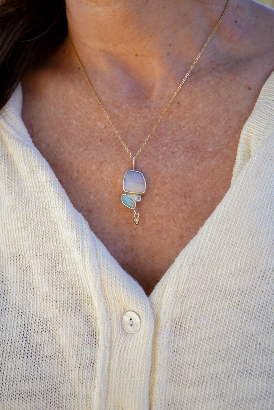A close-up of a person wearing a delicate gold chain necklace with a silver pendant featuring a pale iridescent opals and a small moonstone, paired with a white knit cardigan.
