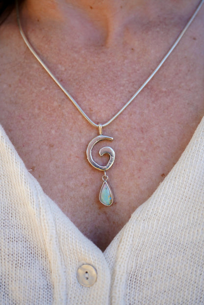 A close-up of a person wearing a silver necklace with a spiral pendant and a dangling  teardrop opal, styled with a cream-colored knit top.