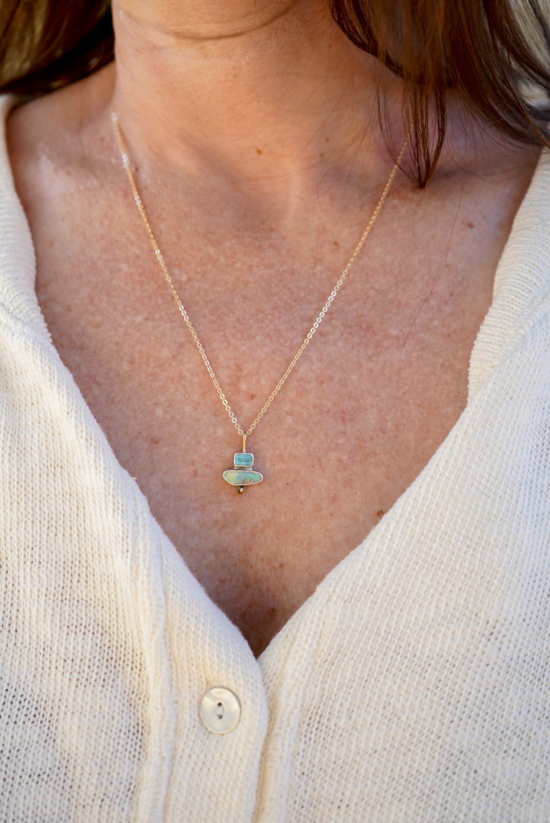 A close-up of a person wearing a delicate gold chain necklace with a small pendant featuring a light blue-green tourmaline stone and opal with a tiny gold accent, styled with a cream-colored knit cardigan.