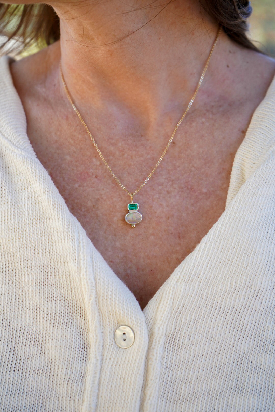 A close-up of a person wearing a delicate gold chain necklace with a small silver pendant featuring a green tourmaline on  top and an opal on the bottom, styled with a cream-colored knit cardigan.