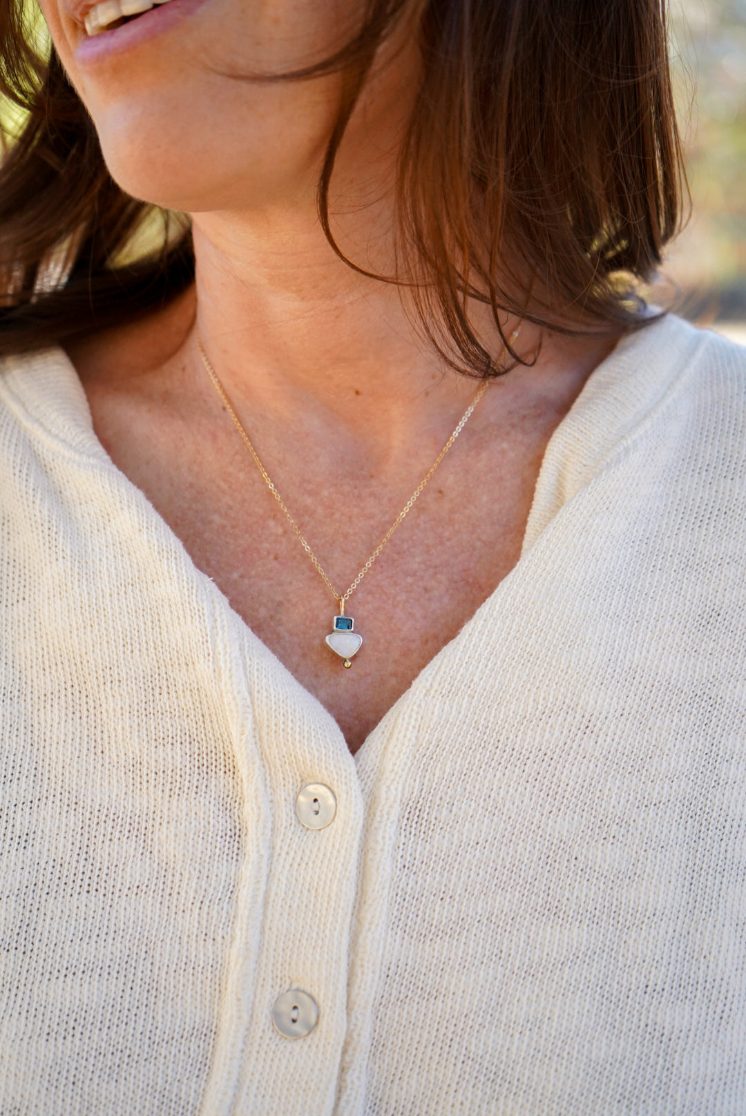 A close-up lifestyle shot of a woman wearing a delicate gold necklace with a dark blue tourmaline and opal pendant, paired with a cream-colored knit cardigan.