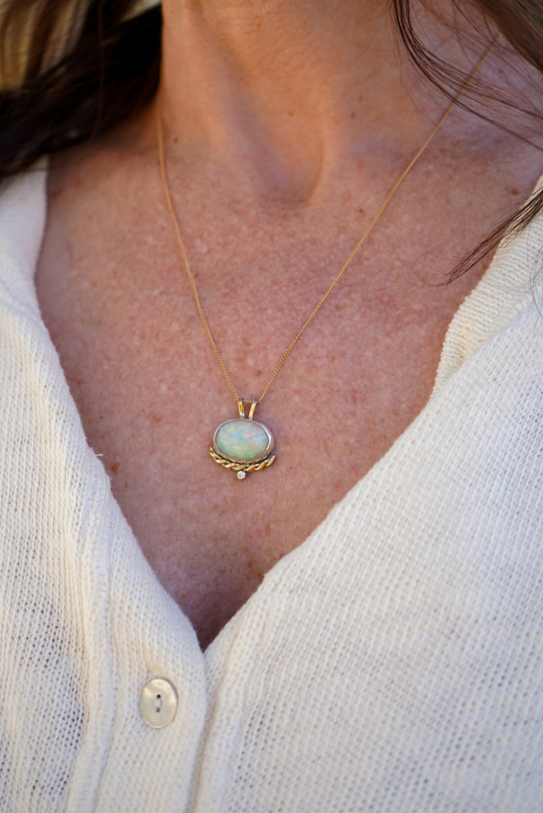 A close-up of a person wearing a delicate gold necklace with an oval light opal pendant, styled with a white knit top.