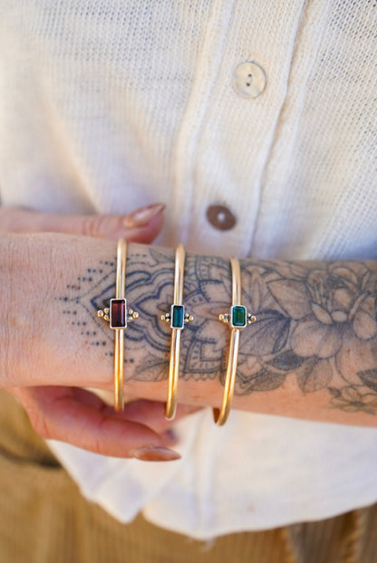 Three gold bangle bracelets with rectangular colored tourmaline stones are shown worn on a person's wrist, with a white button-up shirt visible in the background.