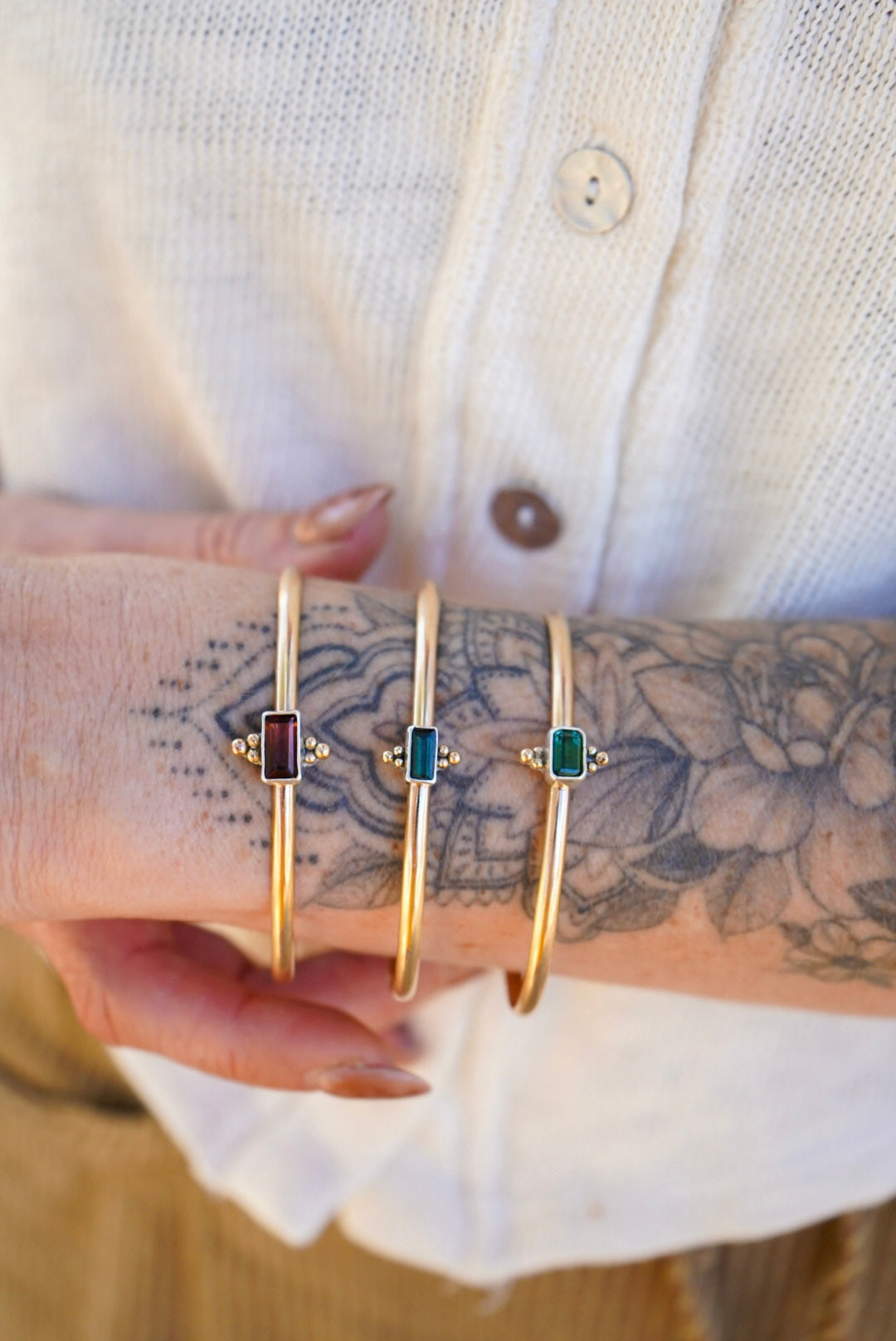 Three gold bangle bracelets with rectangular colored tourmaline stones are shown worn on a person's wrist, with a white button-up shirt visible in the background.