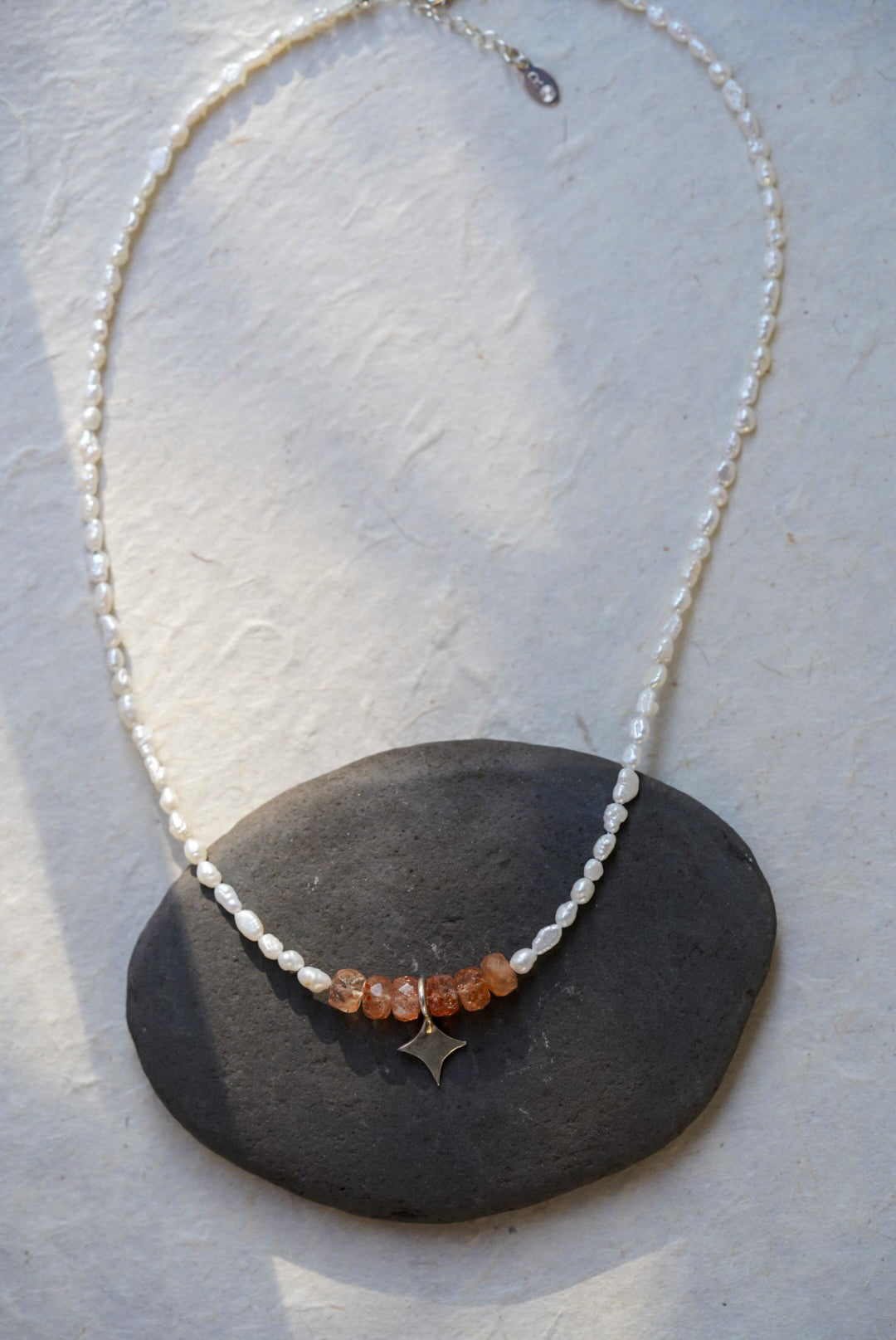 A delicate beaded necklace with white pearl beads, a row of faceted sunstone stones, and a small silver star charm, displayed on a dark stone over a light textured surface.