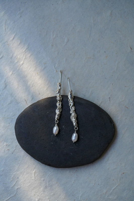 A pair of textured recycled silver dangle earrings and small white pearl-like drops, displayed on a dark oval stone against a light textured wall.