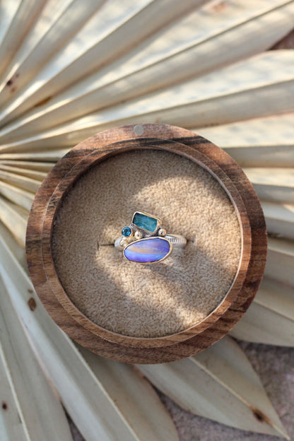 Ring with an opal, tourmaline and blue topaz on a wooden display box with sand, surrounded by dried palm leaves.