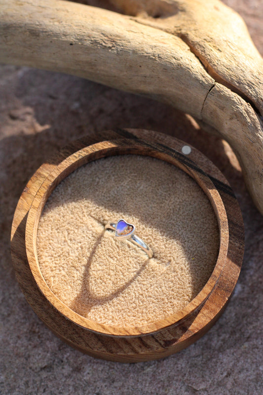 Wooden ring box with a heart shaped  opal ring inside, placed on sand with driftwood.