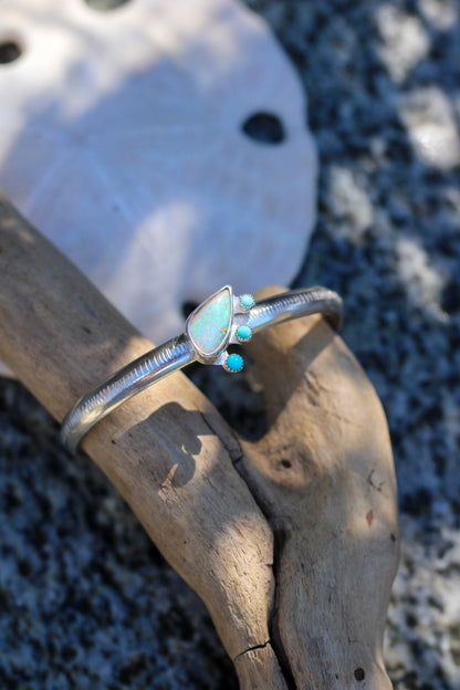Silver cuff with opal and turquoise stones on a piece of driftwood with a sand dollar in the background