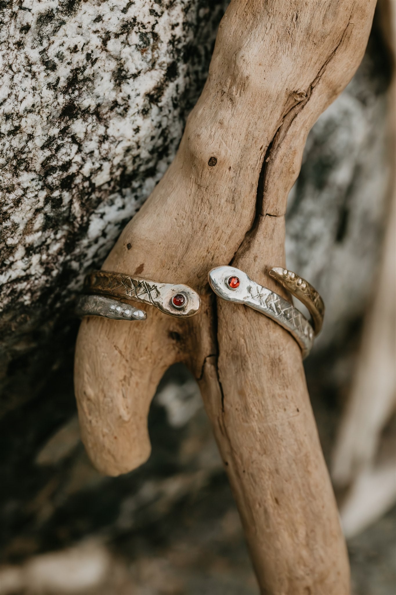 Two silver and bronze cuffs with red stones on a piece of driftwood against a natural background.
