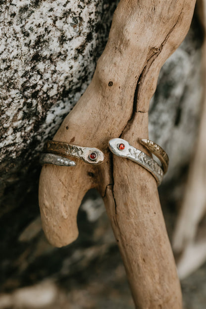 Two silver and bronze cuffs with red stones on a piece of driftwood against a natural background.