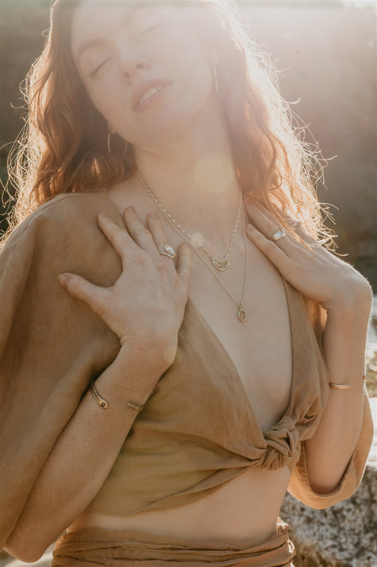 Woman with closed eyes wearing a beige top and gold and silver jewelry against a blurred natural background