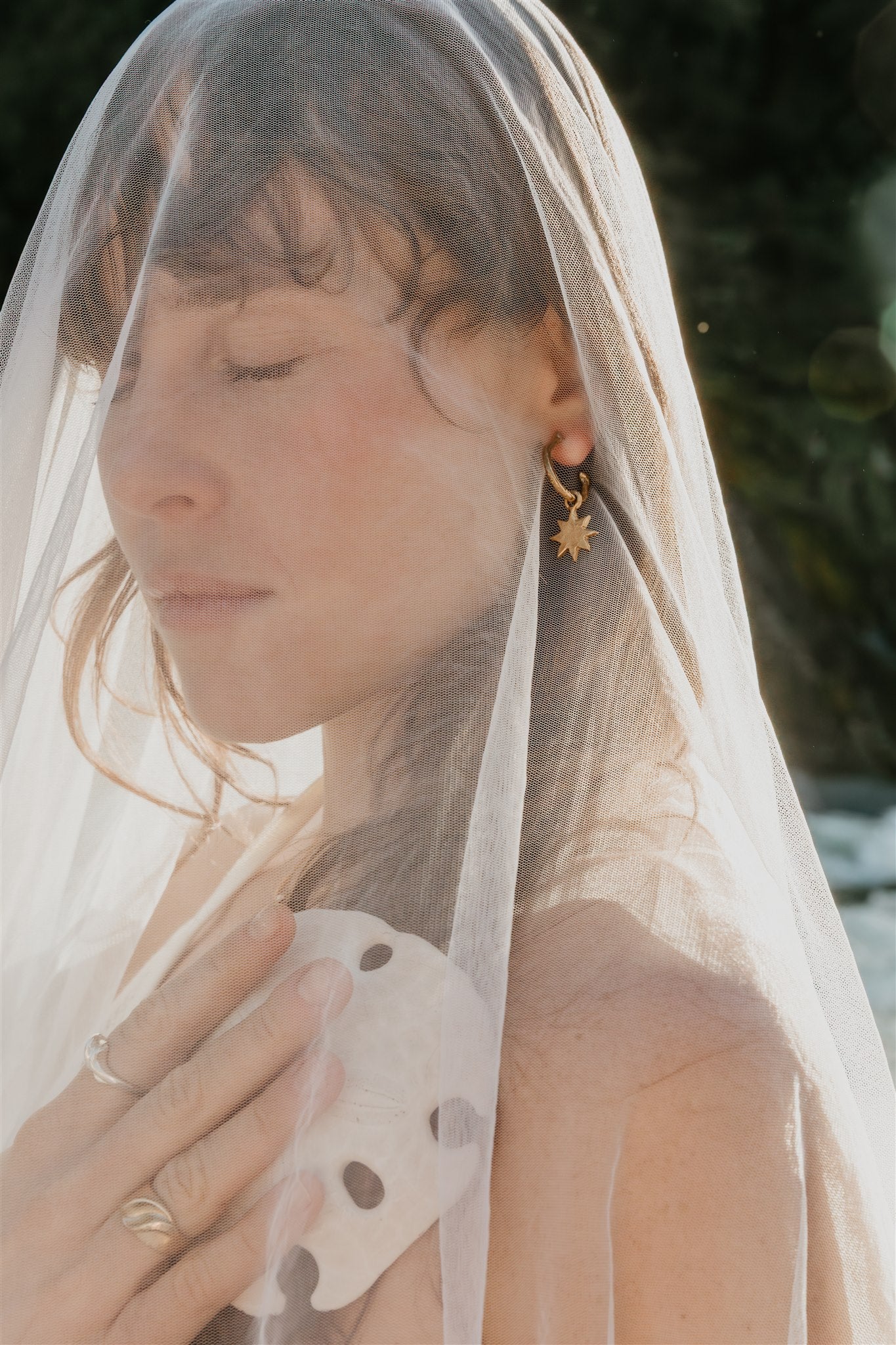 Woman wearing a veil with a blurred natural background, wearing rings and a star earring.