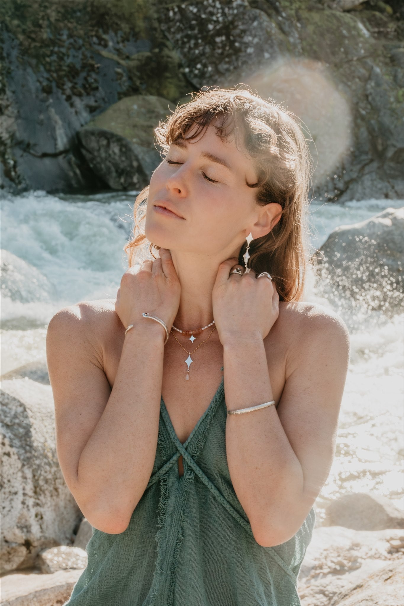 Woman in a green dress standing by a rocky water source, wearing jewelry.