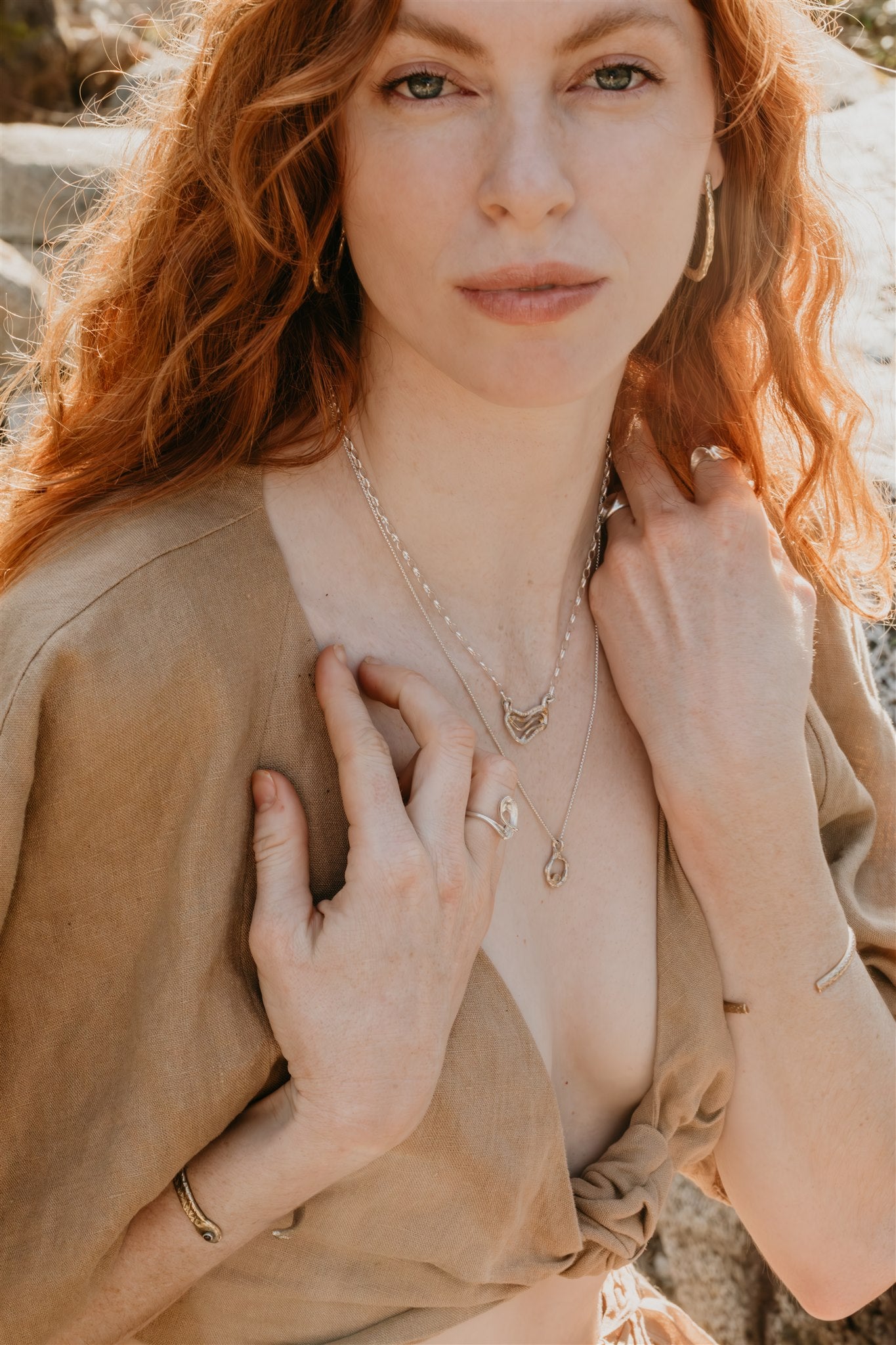 Woman with long hair wearing a beige top and layered necklaces outdoors.